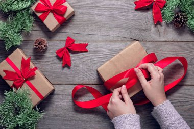 Noel arkaplanı, eller ahşap bir masaya Noel hediyelerini paketliyor, tasarımcı hediye kutularına kırmızı kurdele takıyor.