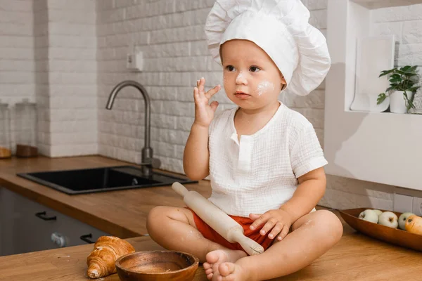 a little cook sits on a table in the kitchen, a 2-year-old boy in a chef\'s hat and flour on his cheeks, mom\'s assistant