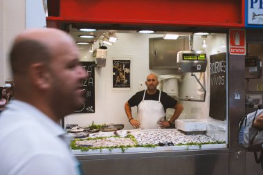 Malaga, İspanya, Temmuz 2019: Tezgahın arkasındaki Atarazanalı adam pazarda balık satıyor, insanlar pazarda dolaşıyor, ürünleri alıyor, seçiyor, şehrin simgelerinden biri olan