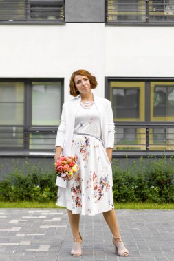 Çiçek buketli beyaz elbiseli bir kadın. Portret. Şehir evinin arka planına karşı.