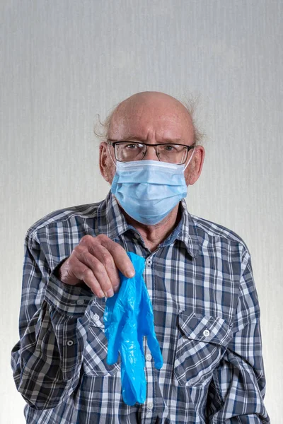 Old bald man with glasses in mask holds the glove medical. Indoors in daylight. Front view.