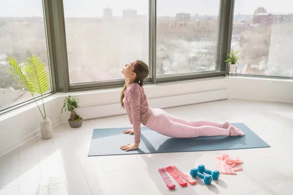 在家练瑜伽的女人在家里做伸展腿的运动，做着向太阳致敬的眼镜蛇的姿势。健身在家里的女孩在早晨的阳光下在公寓客厅里锻炼 — 图库照片