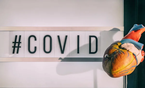 COVID-19 hashtag tecken bredvid medicinsk hjärtmodell visar kardiovaskulär risk för stroke blodpropp med coronavirus infektion. Lightbox skylt text — Stockfoto