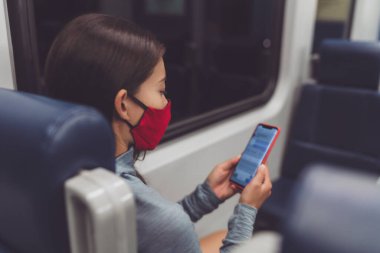 Train commute during corona virus pandemic. Woman passenger using mobile phone wearing face mask sitting in public transport texting online on contact tracing app clipart