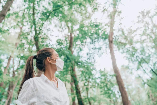 Ormanda yürüyüş yapan kadın, dışarıda yürürken maske takıyor, umutla ağaçlara bakıyor. Temiz hava, sürdürülebilirlik, koronavirüs koruma konsepti için çevre dostu maskeler. — Stok fotoğraf