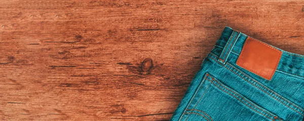 Джинсы брюки на деревянном баннере панорамный фон. Вид сверху на текстуру коричневого дерева и синюю джинсовую одежду для мужчин и женщин с кожаной задней этикеткой — стоковое фото