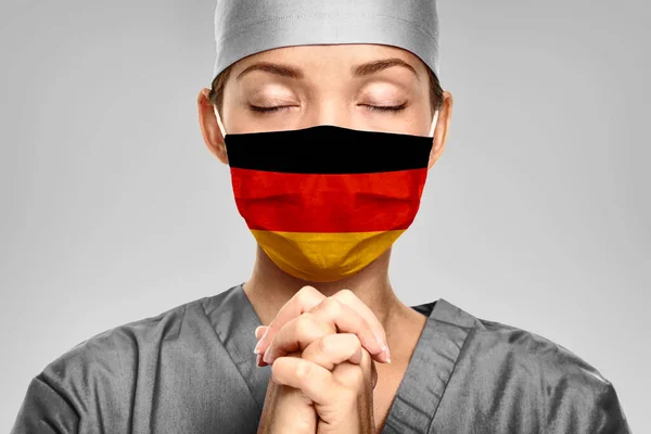 Německý lékař se modlí za pomoc s německou vlajkou na chirurgické masce. Mladá žena lékařské potřeby PPE pomoc v boji proti COVID-19 Coronavirus — Stock fotografie