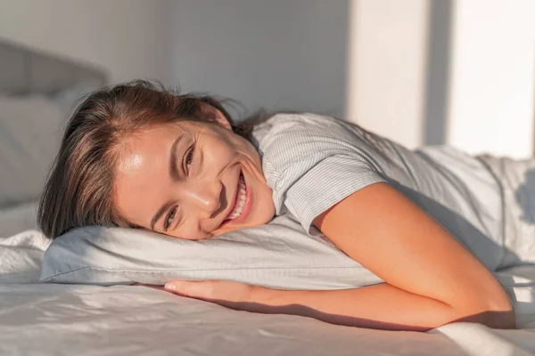 Щасливий дім розслабляє азіатську усміхнену жінку, яка перебуває в ліжку, відпочиваючи в портреті відвертого спальні. Натуральна краса здорового обличчя моделі скінів. Зручний пінопластовий матрац і подушка — стокове фото