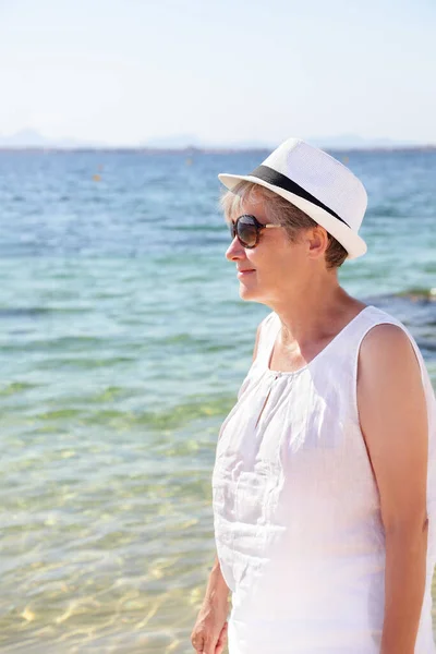 Uśmiechnięta seniorka spacerująca po plaży nad morzem. Starsza pani w stroju plażowym w białym kapeluszu i okularach przeciwsłonecznych. Kobieta na wakacjach wakacyjnych wakacje nad oceanem — Zdjęcie stockowe