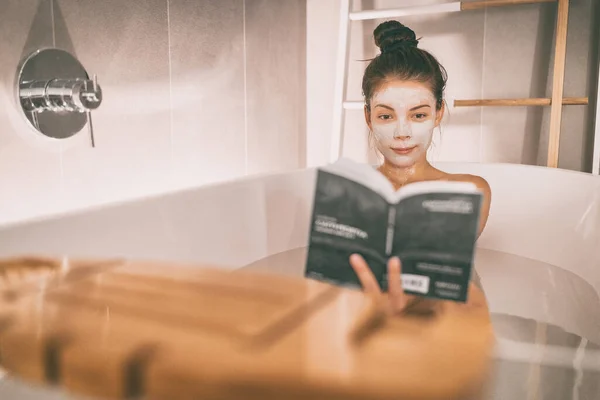 Woman reading book in warm bathtub taking a bath relaxing at home - pamper wellness Asian girl pampering skin care with facial mask treatment — ストック写真