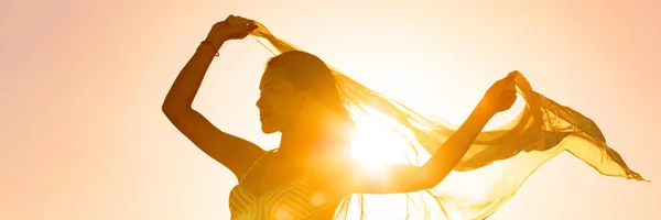 Bella donna felice libera nel bagliore del tramonto sventola sciarpa al vento ballando nel sole banner panoramico. Silhouette femminile signora libertà per il benessere e la vita sana — Foto Stock