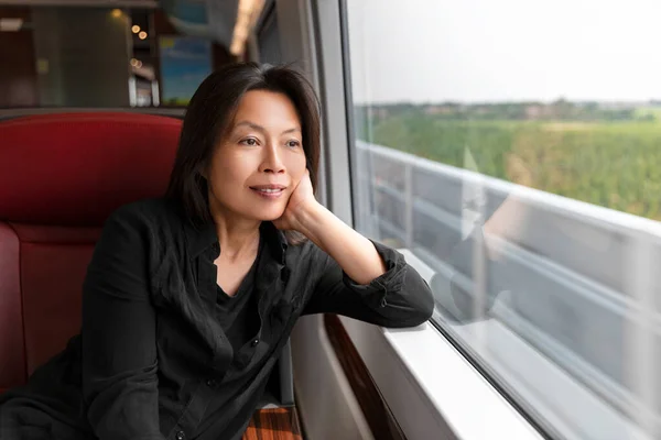 Middle aged Asian woman looking out the window at landscape view sitting on train commute travel lifestyle. Mature chinese passenger traveling smiling relaxing thinking of holiday in contemplation — Stock Photo, Image