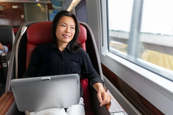 Деловая поездка азиатской женщины, работающей на ноутбуке в поезде пригородного сообщения. Китайская бизнесвумен средних лет улыбается, глядя в окно в созерцании — стоковое фото