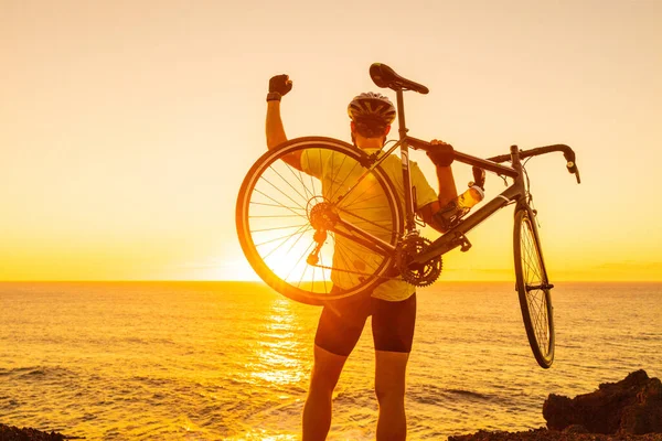 Succès, réussite et concept gagnant avec cycliste homme vélo de route. Heureux athlète professionnel masculin levant les bras levant le vélo par la mer pendant le coucher du soleil acclamant et célébrant au sommet du sommet — Photo