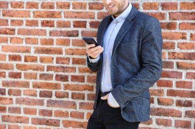 Young casual business man using smartphone in urban city background relaxing on brick wall texting sms on phone app living a modern lifestyle. Closeup of unrecognizable person clipart