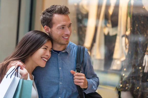 Multiracial couple shopping together looking at clothing in store window walking in city street. Happy asian woman holding shopping bags with caucasian man — Stock Photo, Image