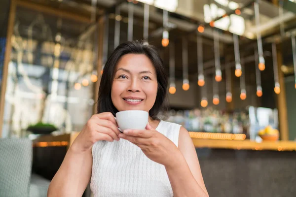 Asian middle aged woman drinking cappuccino coffee at high end fancy cafe. Chinese businesswoman enjoying cup of tea during afternoon at restaurant — Stock Photo, Image
