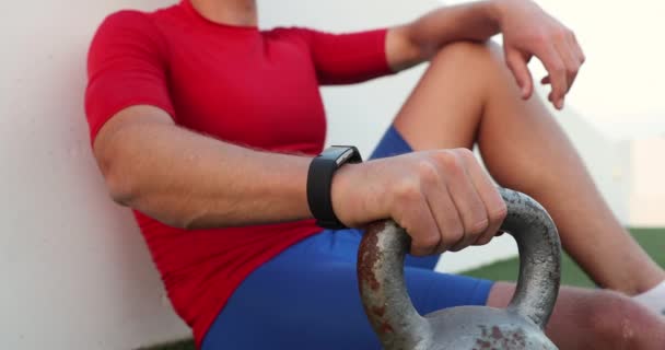 Fitness man op zoek naar fitness activity tracker ontspannen zitten met kettlebell — Stockvideo