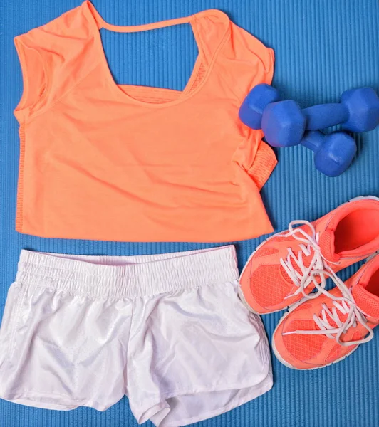 Roupa de ginástica para treino de força fitness layed out em azul tapete de exercício vista superior. T-shirt e sapatos laranja combinando, shorts brancos, pesos halteres. — Fotografia de Stock