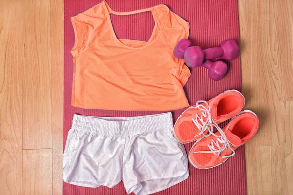 Fitnessbekleidung am Boden für das Krafttraining zu Hause von oben. Orange passende T-Shirts und Laufschuhe, weiße Shorts, rosa Girly-Hanteln auf Matte. — Stockfoto