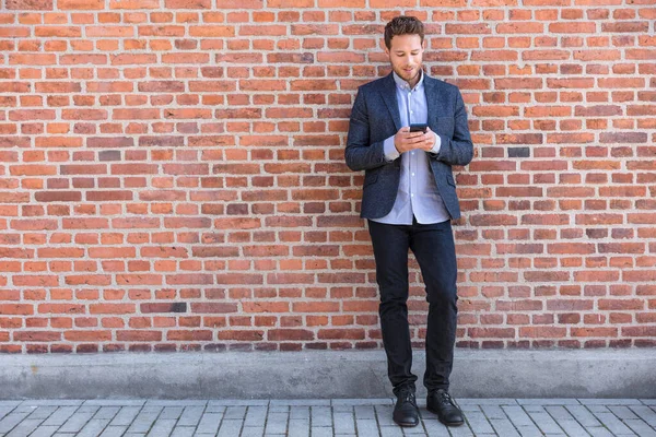 Бизнесмен смс приложение для смс на городской улице на фоне кирпичной стены. Деловой человек держит смартфон в умной повседневной одежде стоя. Городской профессиональный образ жизни — стоковое фото