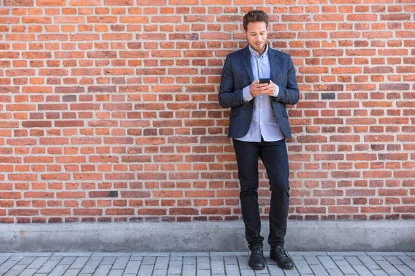 Uomo d'affari sms sms sms telefono app in strada della città su sfondo muro di mattoni. Uomo d'affari che tiene lo smartphone in piedi elegante abbigliamento casual. Urban giovane stile di vita professionale — Foto Stock