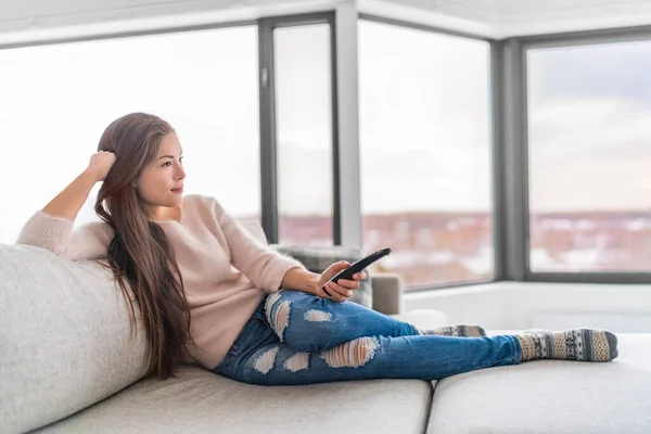 Modern apartmandaki kanepede oturan bir kadın. Genç Asyalı bir kız elinde kumandayla televizyon programının keyfini çıkarıyor. Rahat bir yaşam tarzı. — Stok fotoğraf