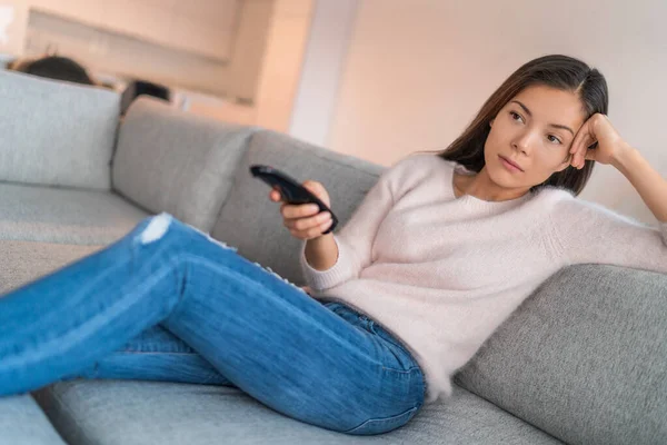 Znudzona kobieta ogląda telewizję sama w mieszkaniu, siedząc na kanapie trzymając zdalnie zamyśloną. Azji dziewczyna zmęczony w domu relaks na kanapie nie mając nic do roboty oprócz oglądania telewizji — Zdjęcie stockowe