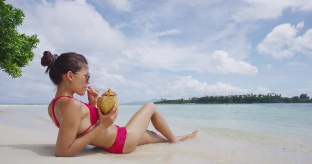 Relaxante mulher tomando banho de sol deitado na areia na praia em biquíni bebendo de coco — Vídeo de Stock