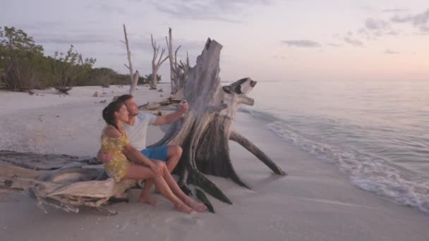 Playa pareja puesta de sol relajarse en vacaciones de luna de miel de verano viajar viendo puesta de sol en Lovers Key, Florida, cerca de Fort Myers Beach, Hombre y mujer tomando fotos de selfie teléfono relajante riendo — Vídeo de stock