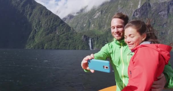 Szczęśliwa para na wakacjach podróżujących biorąc telefon selfie za pomocą smartfona na statku wycieczkowym, Milford Sound, Fiordland National Park, Nowa Zelandia. Wielokulturowa para uśmiecha się śmiejąc SLOW MOTION — Wideo stockowe