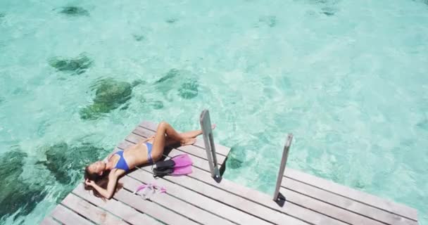 Tropical paraíso viaje vacaciones mujer disfrutando del sol tumbado tomar el sol después de nadar de snorkel de lujo sobre el agua bungalow villa terraza privada en idílica escapada — Vídeo de stock