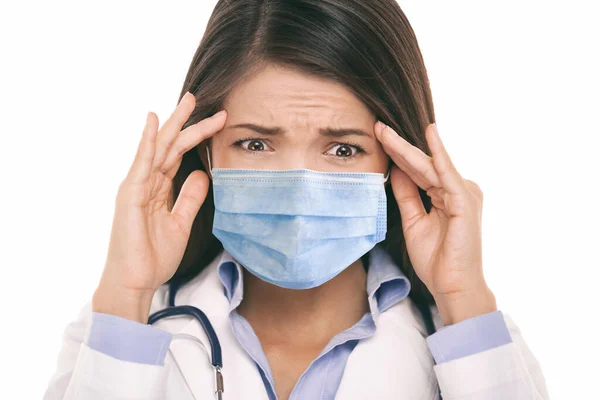 Médico con mascarilla quirúrgica de coronavirus en pánico asustado haciendo una expresión facial estresada divertida sosteniendo la cabeza de dolor de cabeza. Retrato de mujer asiática. — Foto de Stock