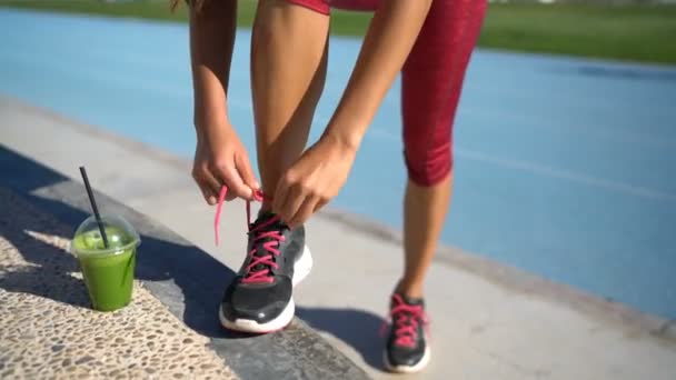 Corredor mujer fitness atando zapatillas de running con batido verde en pista atlética — Vídeo de stock