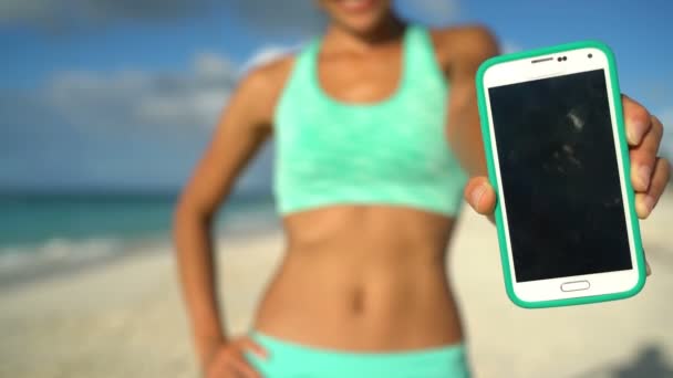 Femme sportive montrant l'application d'entraînement de fitness en cours d'exécution sur l'écran du téléphone sur la plage — Video