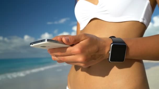 Reloj inteligente y teléfono inteligente en la playa - Mujer en bikini usando tecnología portátil — Vídeos de Stock