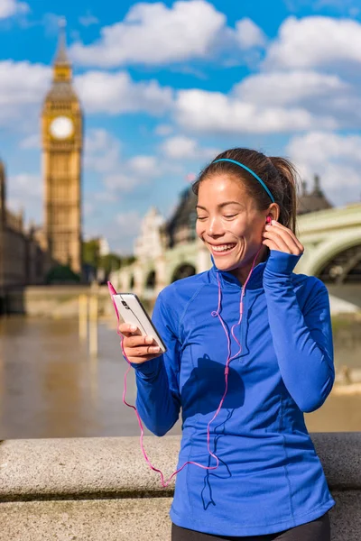 Лондонская бегунья, слушающая музыку на смартфоне возле Биг Бена. Бегущая женщина отдыхает после тренировки в городе. Мбаппе счастливо улыбается на Вестминстерском мосту, Лондон, Англия, Великобритания — стоковое фото