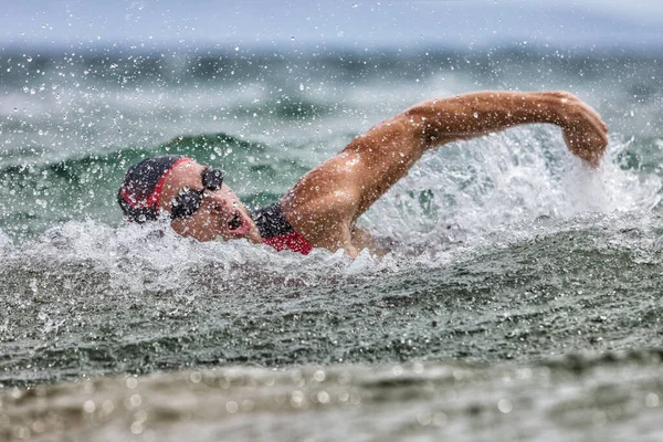 Triathlon nager nageur fatigué nager dans l'océan dans les vagues et la tempête de pluie. Maillot de bain professionnel de triathlon masculin portant un bonnet, des lunettes et un triathlon rouge trois combinaisons d'entraînement pour fer à repasser expirant de l'eau — Photo