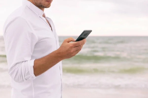 앱을 사용하여 온라인상에서 스마트폰 문자 메시지 sms 를 들고 있는 폰 비즈니스맨. 해변에 사는 사업가가 흰색 바삭바삭 한 셔츠를 입은 채 휴가를 떠나는 길에 대한 정보를 받았습니다. — 스톡 사진