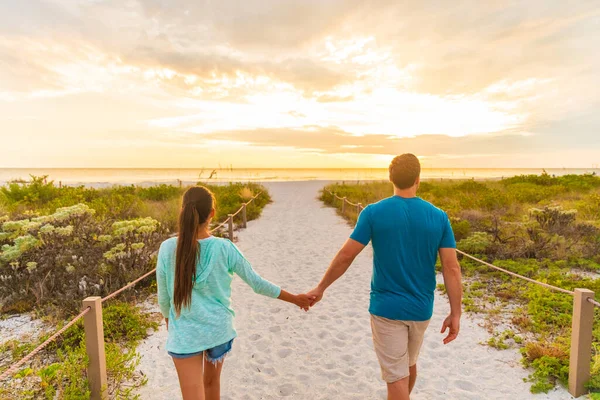 Feliz pareja joven en el amor caminando en romántica noche playa paseo al atardecer. Los amantes de tomarse de la mano en las vacaciones de verano en Florida destino de vacaciones de playa. Gente caminando por detrás — Foto de Stock