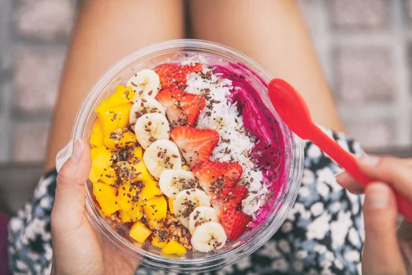 Tazón de pitaya saludable desayuno de frutas. Chica comiendo batido de frutas sentado fuera en el parque para la pausa para el almuerzo. comida vegana primer plano. Puré de Dragonfruit, plátanos, fresas, mangos, coco, semillas de lino —  Fotos de Stock