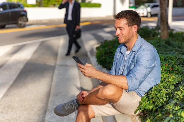 Telefon komórkowy szczęśliwy młody człowiek smsy SMS na smartfonie czeka na spotkanie na ulicy miasta. Miejski styl życia ludzi na świeżym powietrzu w lecie — Zdjęcie stockowe