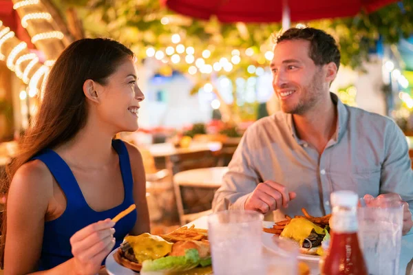 Pareja comiendo hamburguesas en restaurante al aire libre terraza turistas felices en vacaciones de verano. Florida viajan personas que comen alimentos por la noche durante las vacaciones en Miami. Asiático caucásico interracial jóvenes adultos —  Fotos de Stock