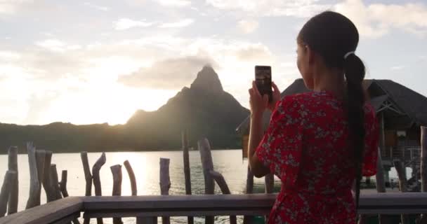 Luxe reisvakantie. Toeristische vrouw met behulp van de telefoon om foto 's te maken bij zonsondergang uitzicht op de oceaan en het berglandschap op Bora Bora, Frans-Polynesië, Tahiti. Luxe woonresort levensstijl — Stockvideo