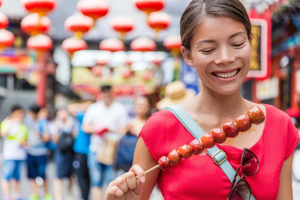 中国の女性ビンタンフル、北京から伝統的な中国料理のスナックを食べる。果物キャンディースティック王府井通りの屋外市場の場所で販売されます。竹の棒の上に砂糖漬けの果物 — ストック写真