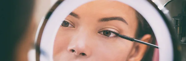 Vrouw zet mascara make-up in spiegel banner thuis badkamer ochtend routine. Mooi Aziatisch meisje klaar voor het aanbrengen van oog make-up met borstel. Close-up op het oog in reflectie — Stockfoto