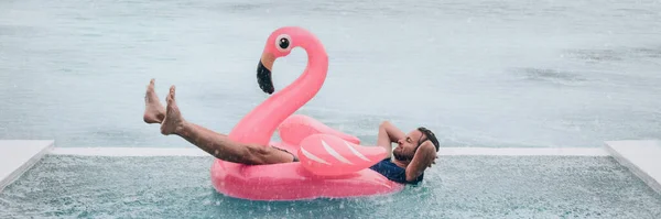 Happy man relaxing in swimming pool flamingo float despite bad rain weather. Travel summer vacation banner. — Stock fotografie