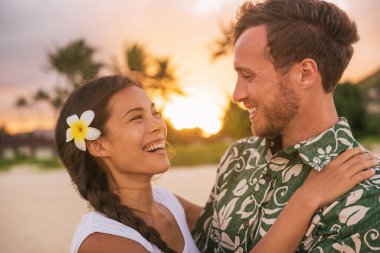 Happy couple lovers in love on romantic sunset beach vacation in Hawaii travel. Asian woman hugging Caucasian man smiling, interracial relationship. Newlyweds honeymoon or wedding on Tahiti beach. clipart