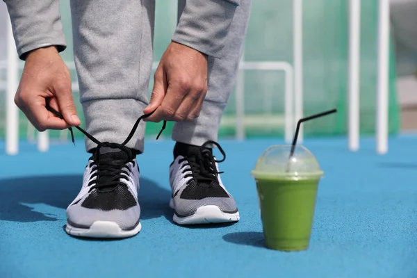 Spor salonundaki adam spor ayakkabılarını yeşil smoothie detoks içeceğiyle bağlamaya hazırlanıyor. Plastik bardak sebze suyuyla koşan spor ayakkabılara yakın çekim. Spor etkin yaşam tarzı — Stok fotoğraf