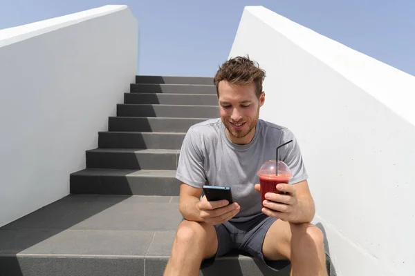Saludable smoothie beber hombre usando aplicación de teléfono en jogging descanso con la mañana jugo de remolacha roja sentado en escaleras al aire libre — Foto de Stock